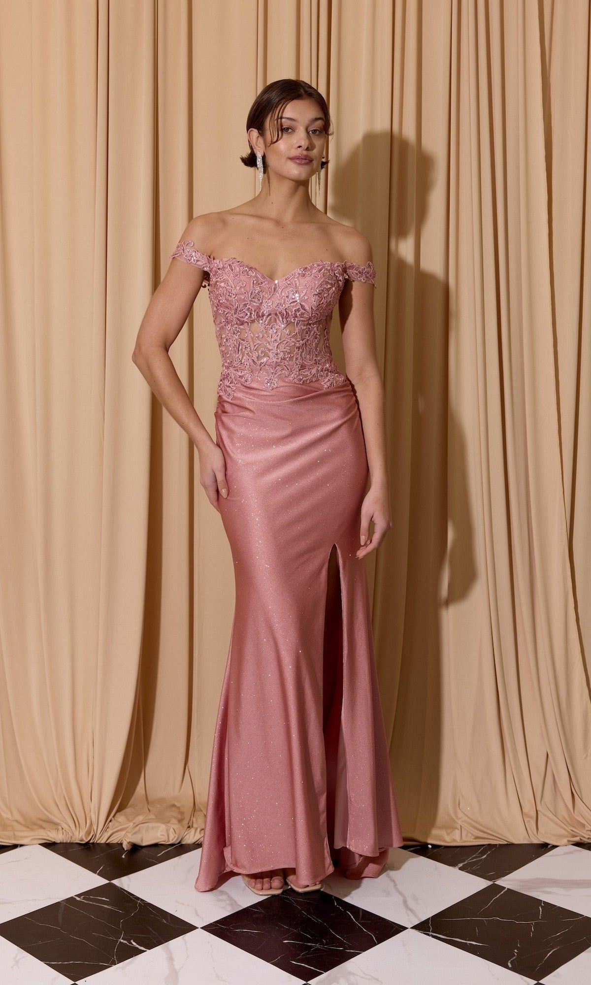 A woman wearing the Eureka EK173 Long Prom Dress with a lace bodice and slit skirt stands on a black and white tiled floor in front of beige curtains.